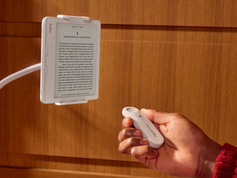 Eine weiße Kobo Remote wird in der rechten Hand einer Person gezeigt, die auf einen weißen Kobo Libra Colour eReader zeigt, der auf einem biegbaren Ständer steht.