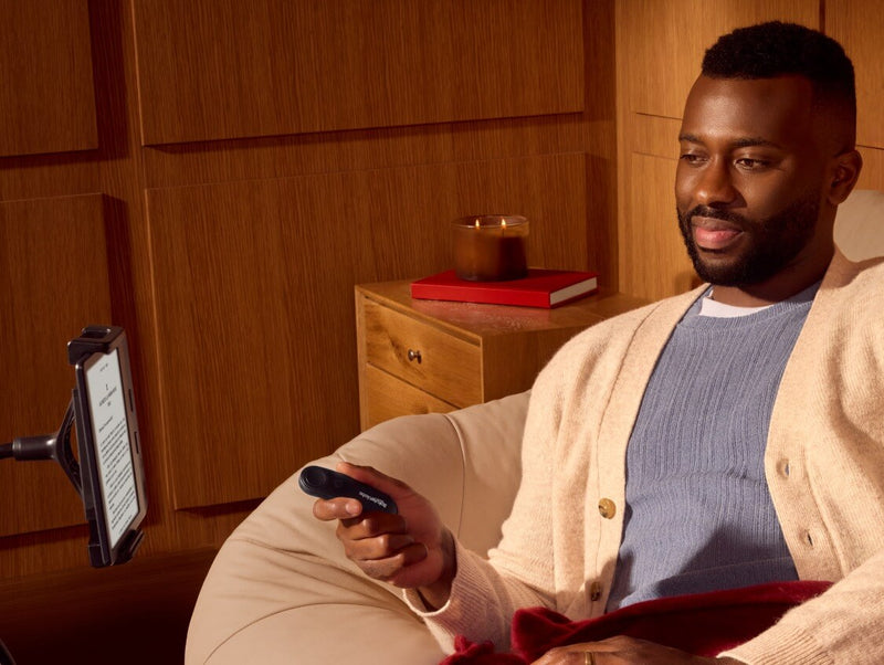 Un hombre, sentado cómodamente, sostiene un Kobo Remote en su mano derecha mientras mira un Kobo eReader sobre un soporte flexible.