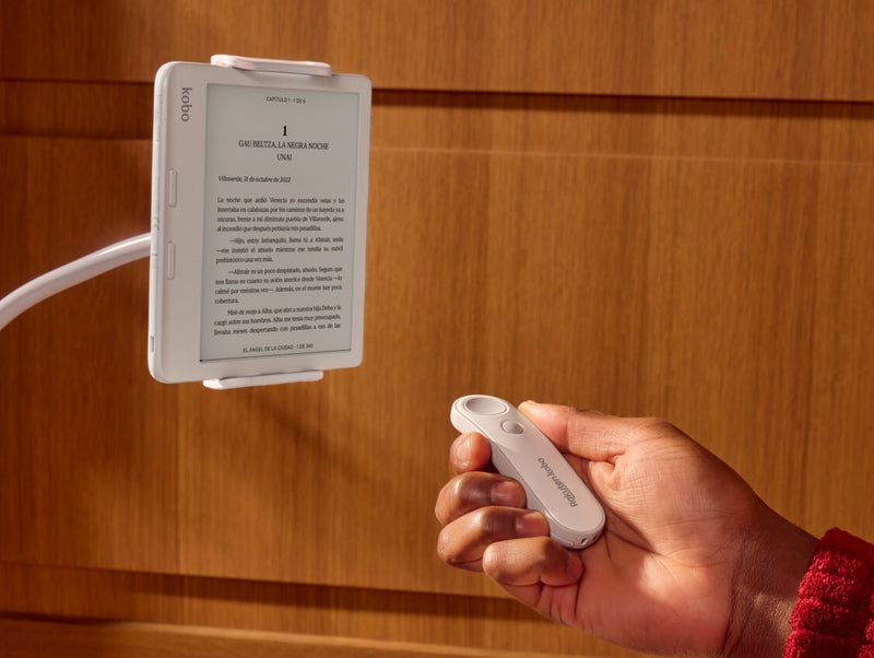 Se muestra un Kobo Remote blanco en la mano derecha de una persona, apuntando hacia un eReader Kobo Libra Colour blanco sobre un soporte flexible.