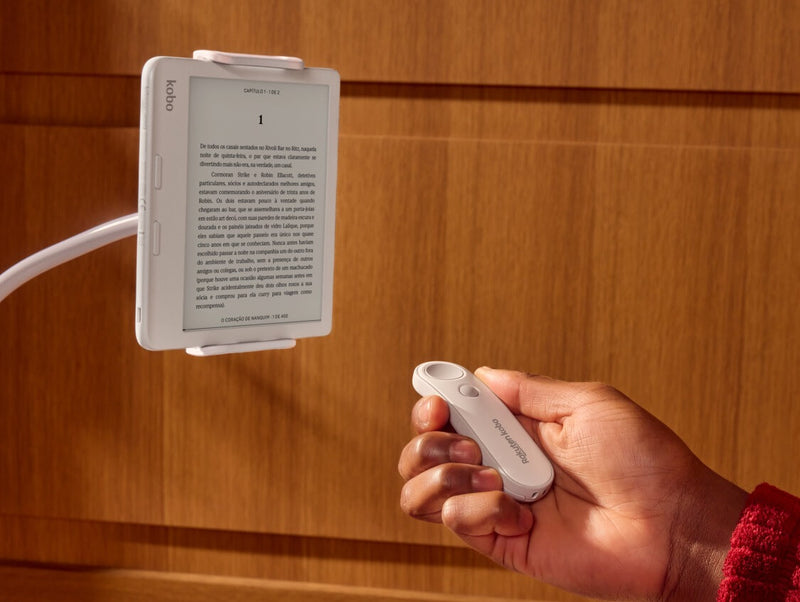 Um Kobo Remote branco é mostrado na mão direita de alguém, apontando para um eReader Kobo Libra Colour branco em um suporte ajustável.